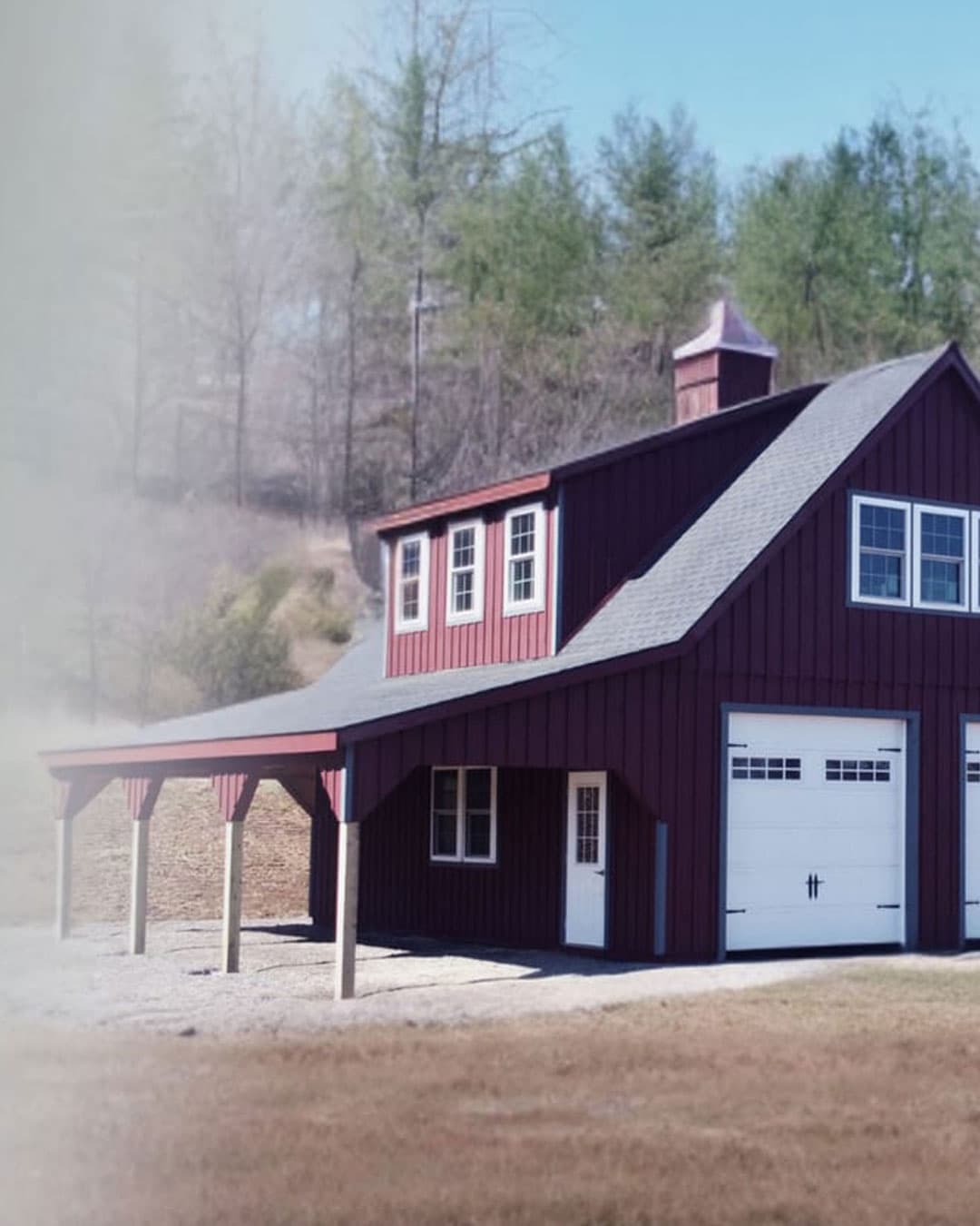 Custom two-story burgundy barn-garage with lean-to, cupola, and dual carriage-style garage doors — Shenandoah-Sheds-built for a Virginia property